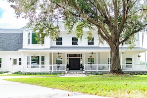 A two-story white house with a porch, a large tree in front, and a well-maintained lawn.