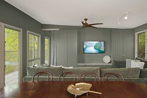 A dining area with a wooden table, a television mounted on the wall, and a sofa with a view of greenery outside.