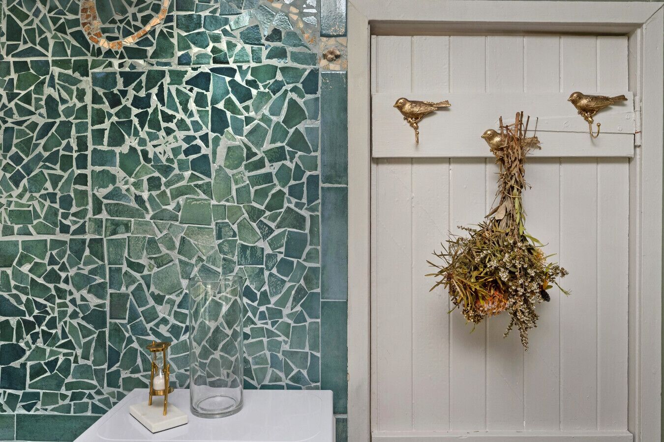 A bathroom with a mosaic tile wall, a white door with bird hooks, and a clear glass vase on a white surface.