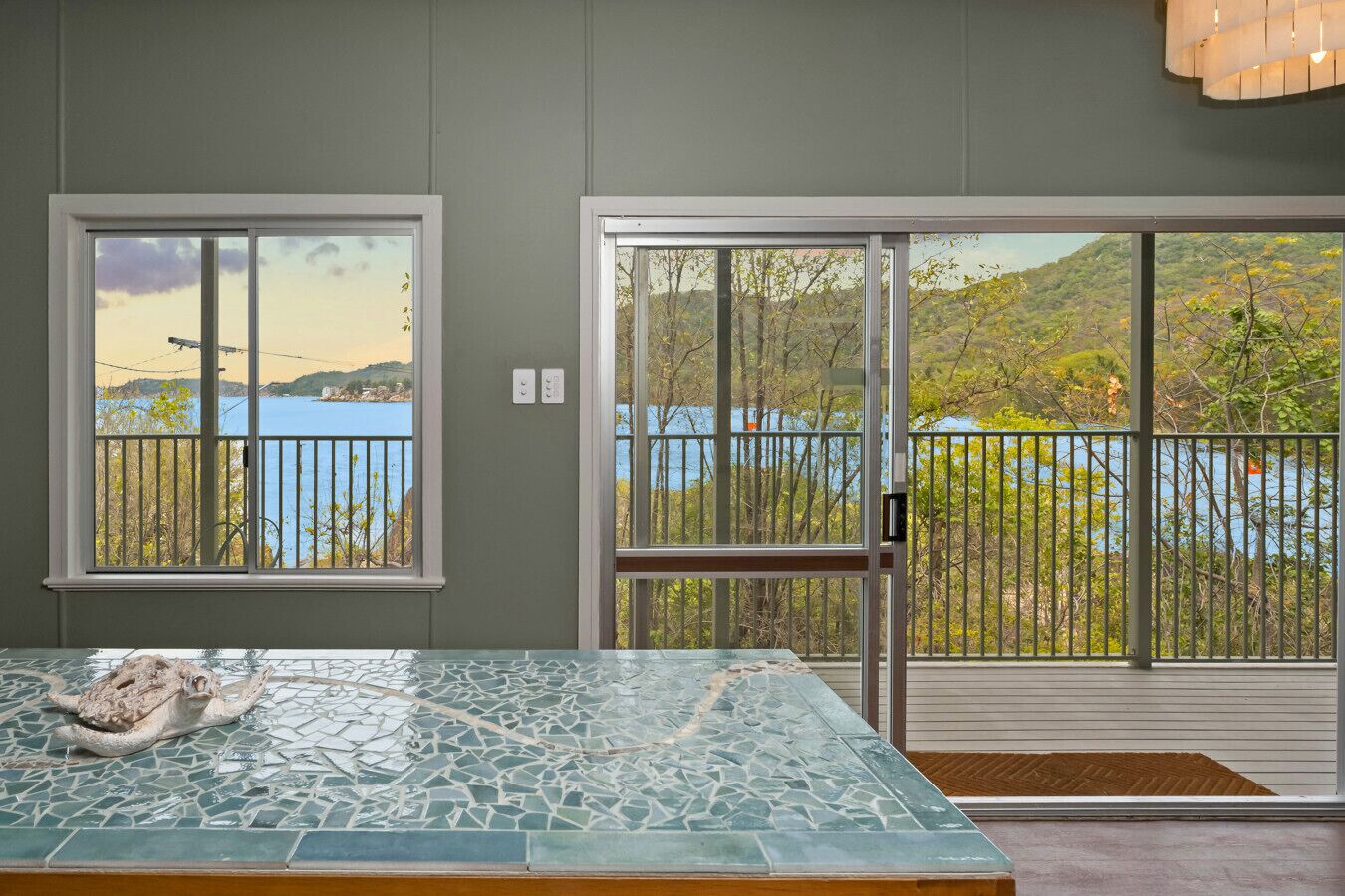 Una habitación con una mesa de vidrio, vista a un lago a través de ventanas amplias y un balcón con barandilla.