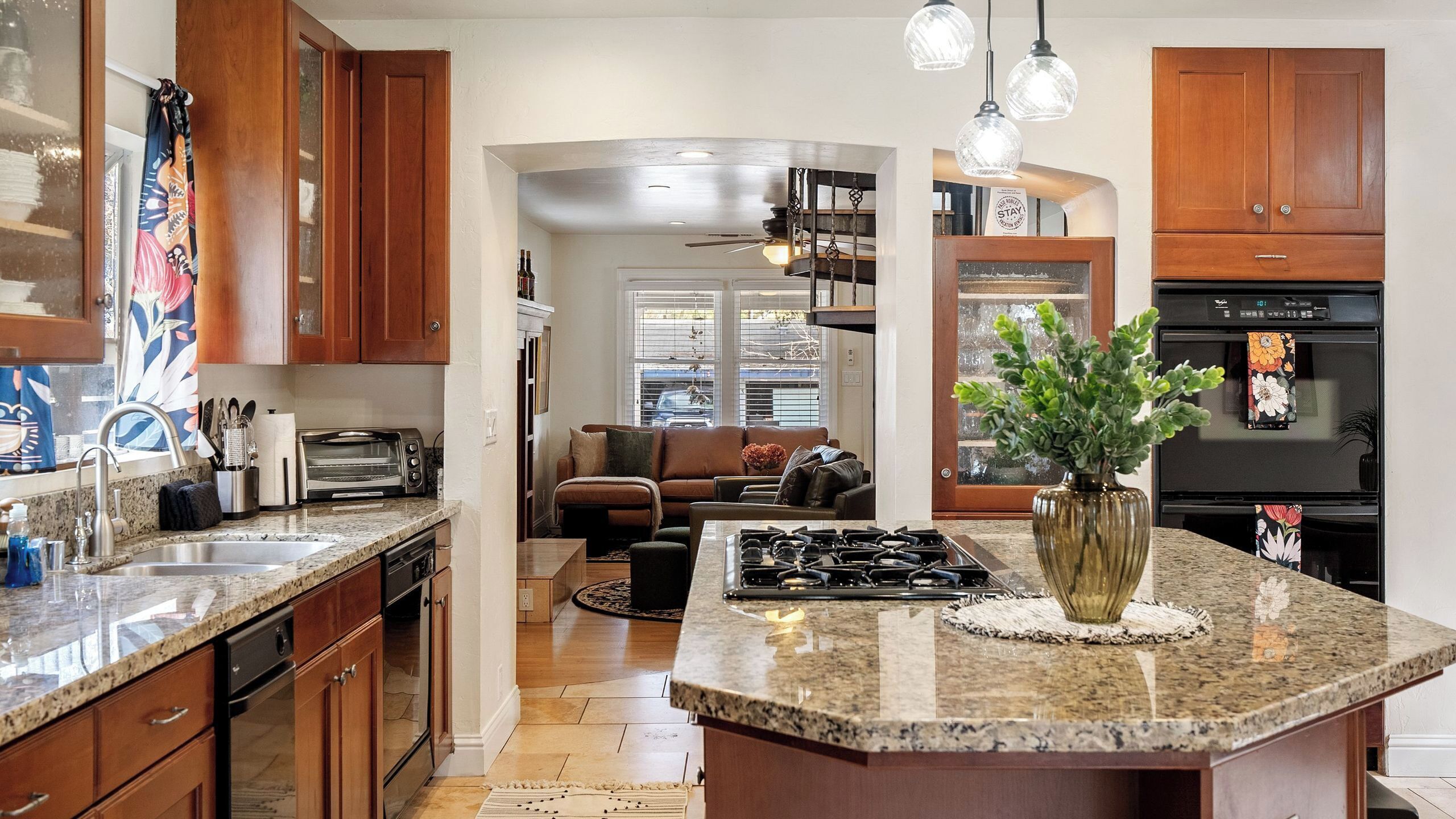 Cocina con encimeras de granito, gabinetes de madera, una isla central y un comedor con sofá y mesa de centro.