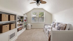 Una habitación con techo inclinado, una ventana con persianas, una estantería, una cama blanca y un cubo de basura pequeño y blanco.