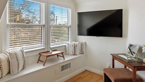 Una habitación con un ventanal grande, un televisor de pantalla plana en la pared, una mesita de madera y un banco con cojines.