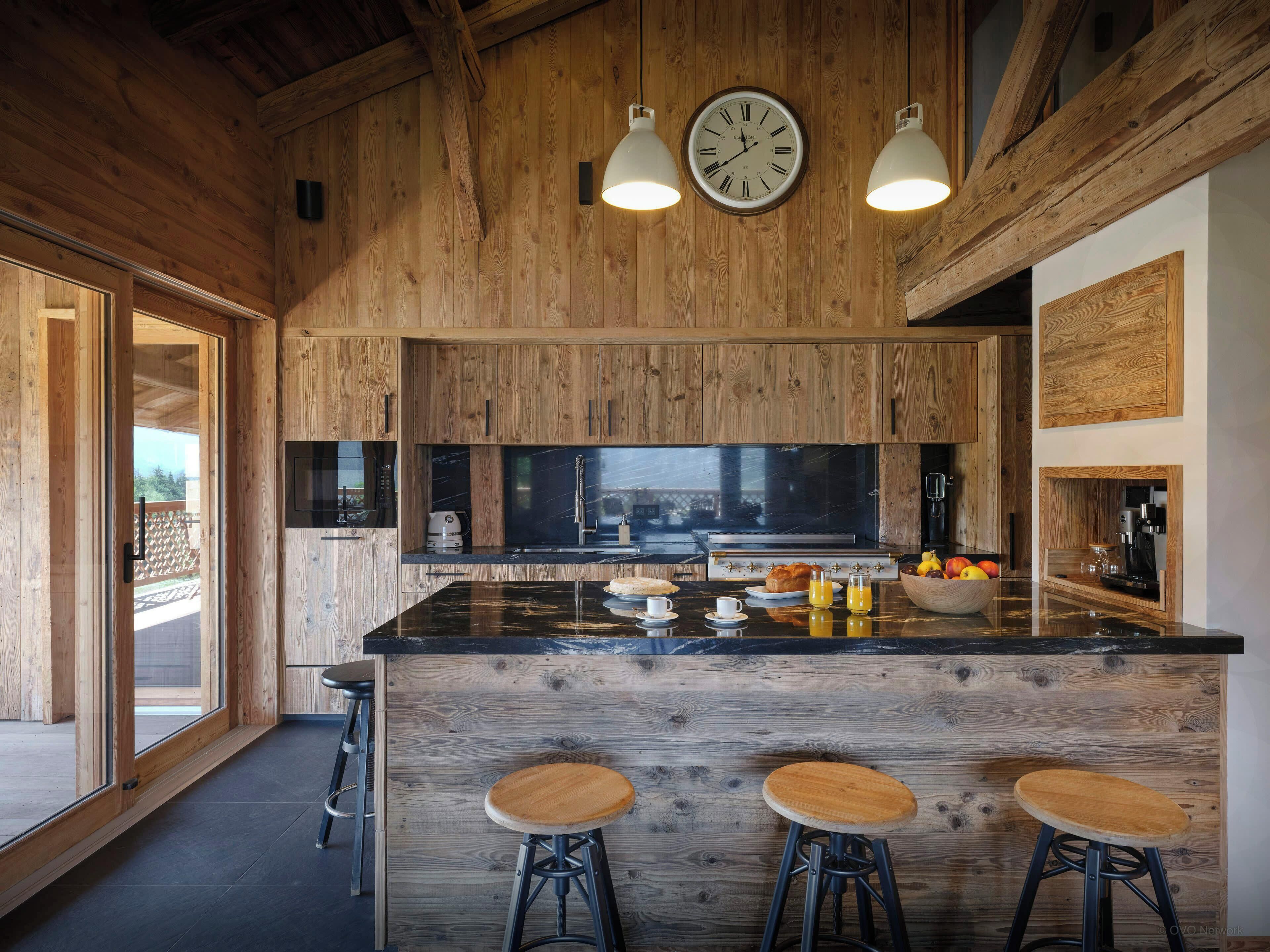 A modern kitchen with wooden paneling, a large island with stools, and a clock above the island.
