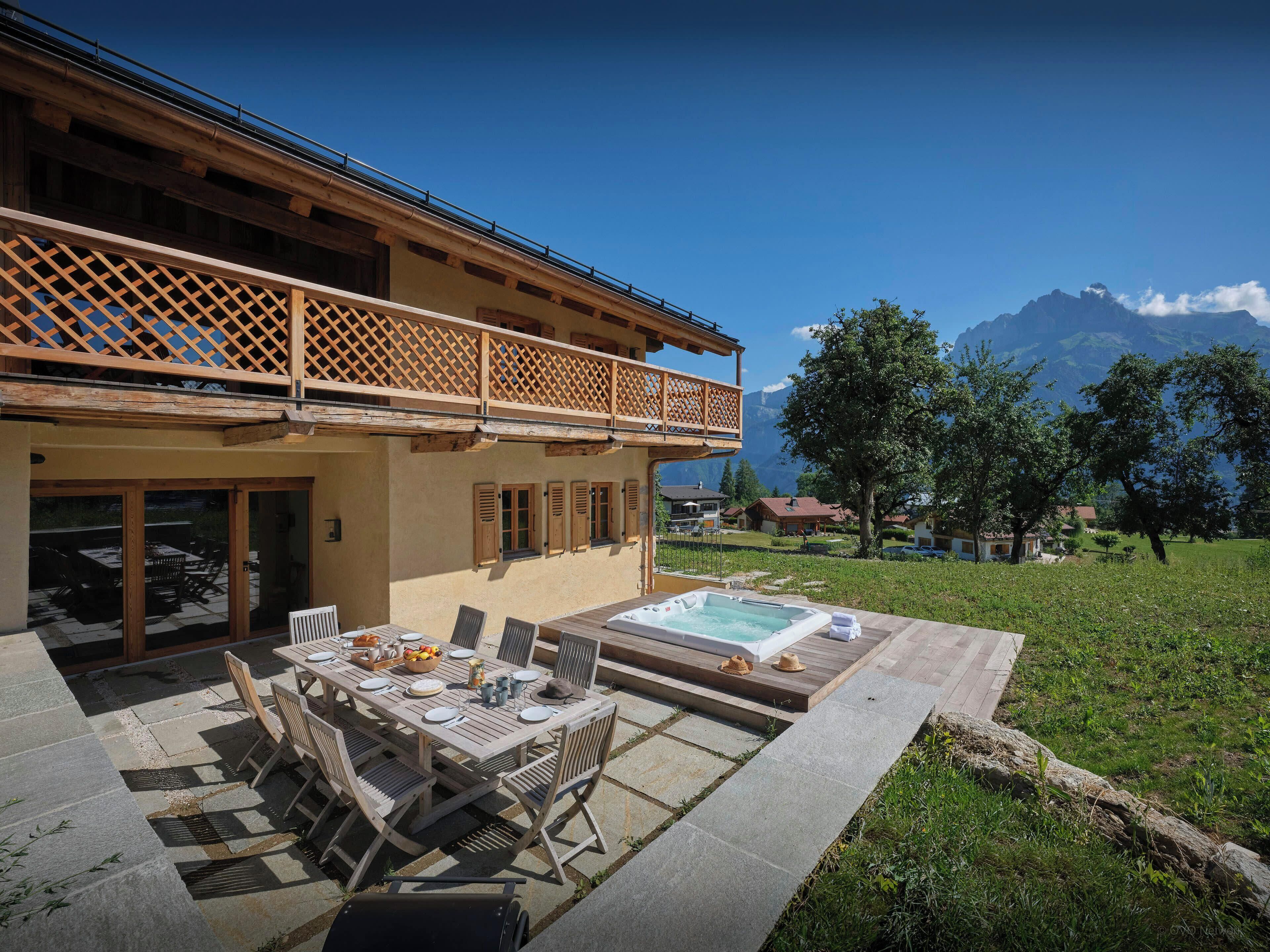 A chalet with a wooden balcony, a hot tub, and a dining area with outdoor furniture.