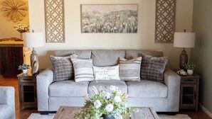 A living room with a grey sofa, multiple cushions, a coffee table with flowers, and decorative items on side tables. - Bella Vista Farmhouse by Avantstay River Views + Fire Pit + BBQ Paso Robles Wine Country (Paso Robles)