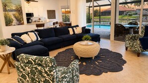 A spacious living room with a dark blue sofa set, a round woven coffee table, and patterned armchairs. A large window offers a view of a swimming pool and outdoor furniture. - Gullaby Getaway - Pool, Wide Canal, Sleeps 7 (Cape Coral)