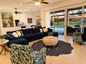 A spacious living room with a dark blue sofa set, a round woven coffee table, and patterned armchairs. A large window offers a view of a swimming pool and outdoor furniture. - Gullaby Getaway - Pool, Wide Canal, Sleeps 7 (Cape Coral)
