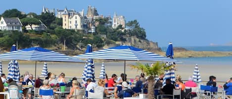 A seaside restaurant with outdoor seating and umbrellas, overlooking a beach and historic buildings.