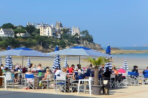 A seaside restaurant with outdoor seating and umbrellas, overlooking a beach and historic buildings. - La Petite Quinoise - PLOUER SUR RANCE (PLOUER SUR RANCE)