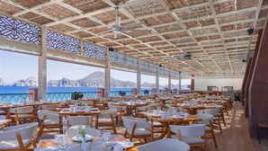 Een ruime eetruimte met gedekte tafels voor gasten, grote ramen met uitzicht op zee en een plafond met een geometrisch patroon.