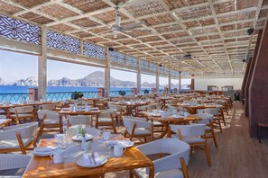 A spacious dining area with tables set for guests, large windows offering a view of the sea, and a ceiling with a geometric pattern. - Luxury Pool-View Studio at 5-Star Villa del Arco– Steps to Medano Beach! (cabo san lucas)