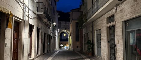 Una calle empedrada bordeada por edificios con balcones y toldos, que lleva a un arco distante.