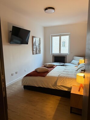 Une chambre à coucher avec un lit, des lampes de chevet, un téléviseur fixé au mur, une fenêtre donnant sur un beau paysage et un radiateur.