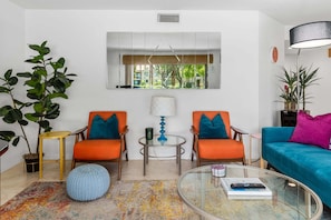 Ein modernes Wohnzimmer mit zwei orangenen Sesseln, einem blauen Sofa, einem runden Glastisch und einer großen Pflanze.
