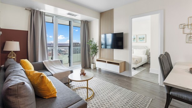 A modern hotel room with a sofa, a dining table, a TV, and a balcony with a city view.