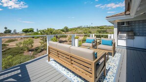 Eine Terrasse mit einer Couch und Sesseln, einem Grill und Blick auf eine hügelige Landschaft.