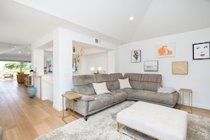 Ein modernes Wohnzimmer mit einem grauen Ecksofa, einem weißen Hocker und einem flauschigen Teppich. An den Wänden sind Kunstwerke angebracht und eine Treppe führt auf eine weitere Ebene.