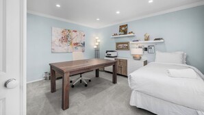Ein Schlafzimmer mit Bett, Schreibtisch, Stuhl, Lampe und Kunstwerken an der Wand.