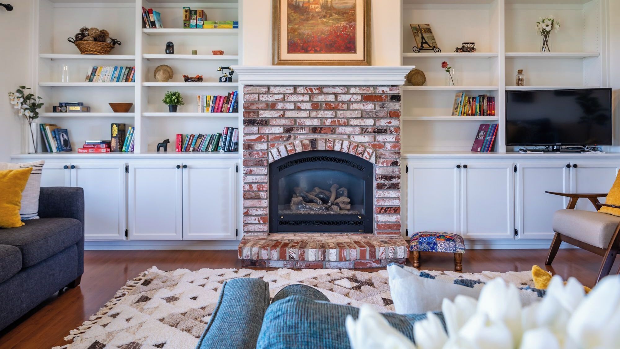 Una sala de estar con chimenea de ladrillo, estanterías blancas con libros y adornos, un sofá y un televisor.