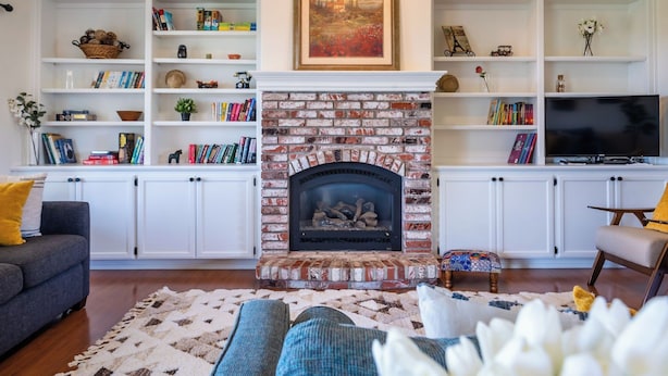 Uma sala de estar com lareira de tijolos, estantes brancas com livros e objetos decorativos, um sofá e uma televisão.