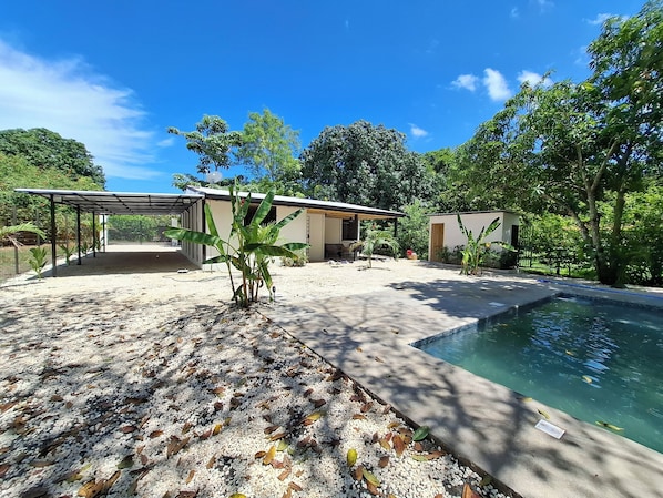Área externa coberta com piscina, cercada por vegetação e um edifício com varanda.