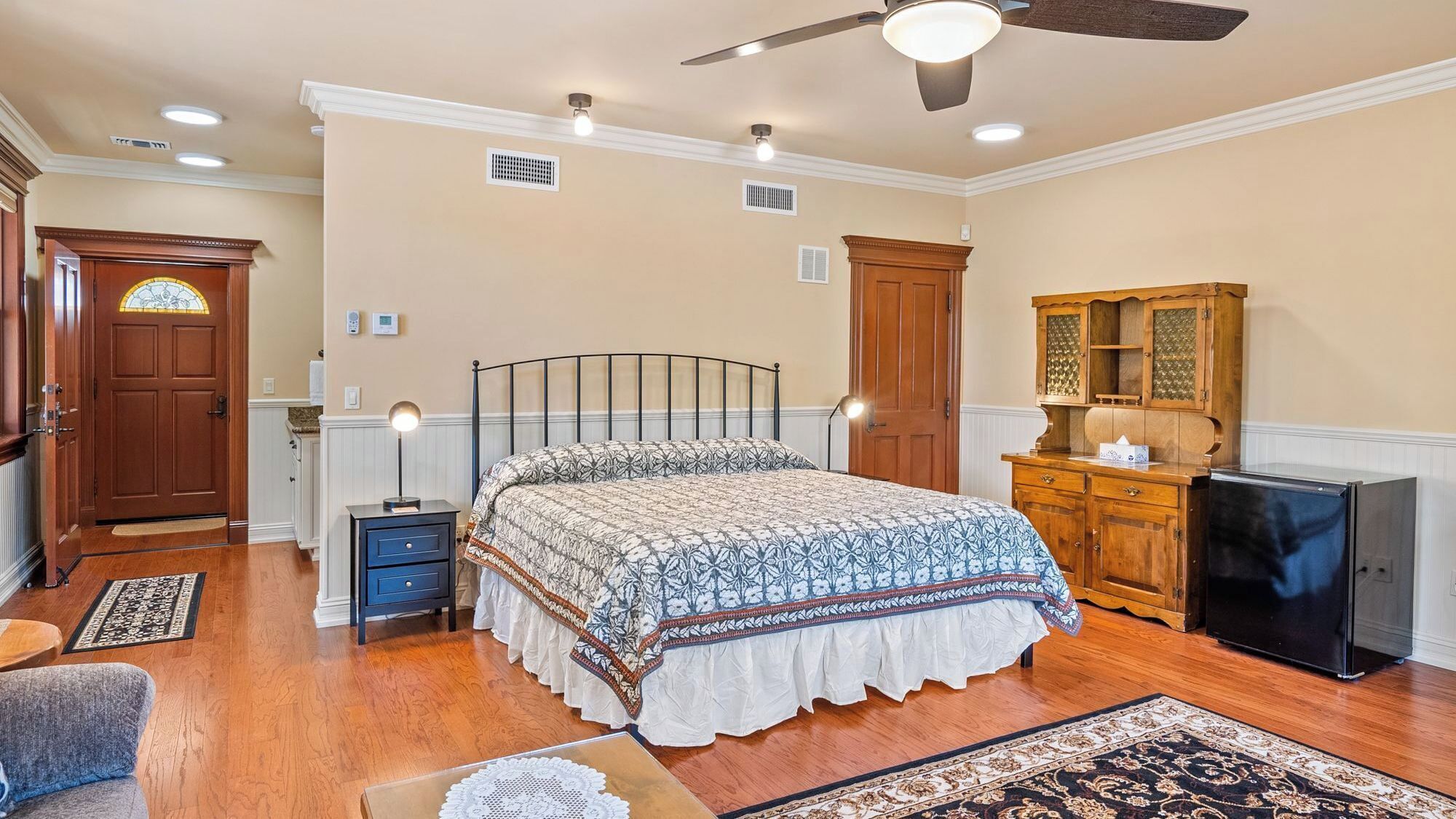 A bedroom with a bed, bedside tables, a wardrobe, a desk, a refrigerator, and a ceiling fan.