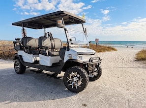 Ein Golfmobil steht auf einem sandigen Pfad, im Hintergrund liegen Strand und Meer.