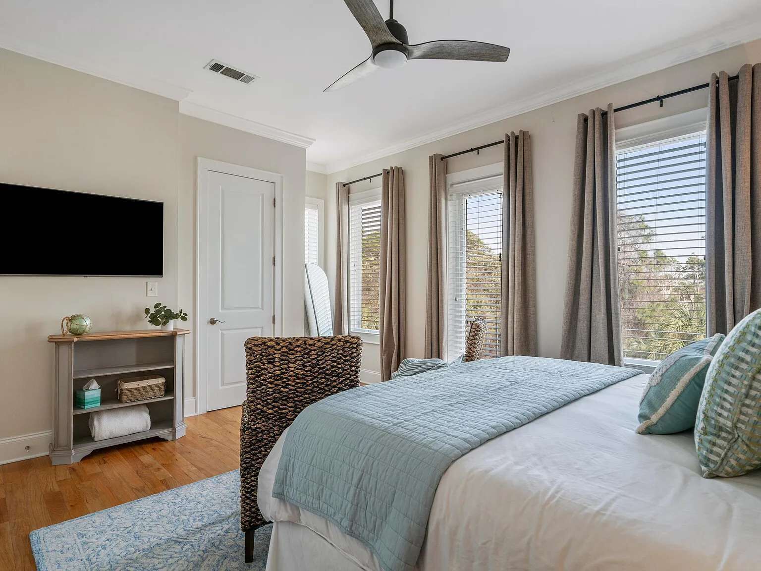 Quarto com cama, ventoinha no teto, televisão fixada na parede, janela com cortinas e prateleira com objetos.