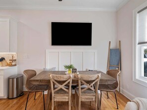 A dining area with a wooden table and four wicker chairs, a television mounted on the wall, and a window with blinds. - Amazing Rooftop Views | 2 Min Walk to Beach (Santa Rosa Beach)