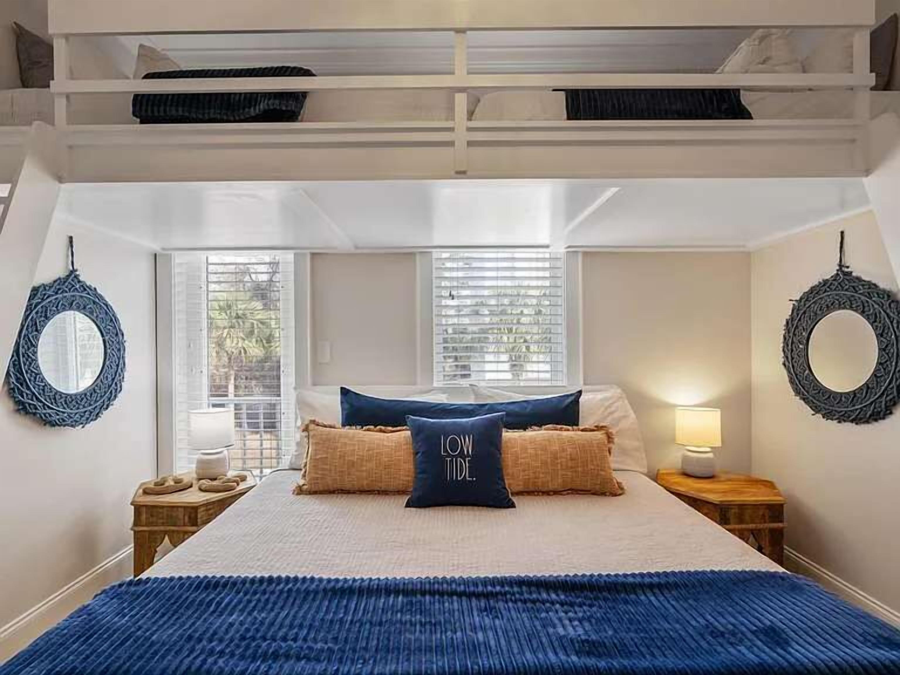 Un dormitorio con literas, una cama grande con una manta azul, dos mesitas de noche con lámparas y un espejo circular en la pared.
