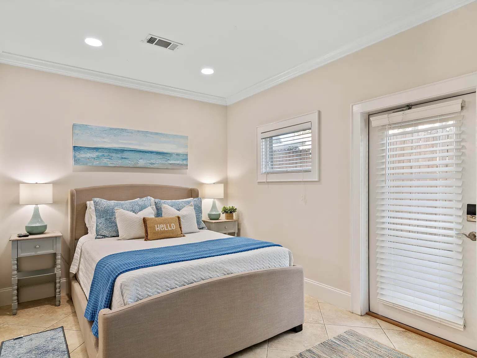 A bedroom with a bed, bedside tables, a window with blinds, a painting on the wall, and a door with a window.