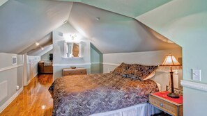 Ein Schlafzimmer mit schrägem Dach, einem Bett mit gemusterten Betttextilien, einem Nachttisch, einer Lampe und einem Spiegel an der Wand.