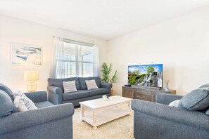 Ein Wohnzimmer mit grauem Sofa, einem Couchtisch, einem Fernseher auf einem Schrank und einem Fenster mit Jalousien.