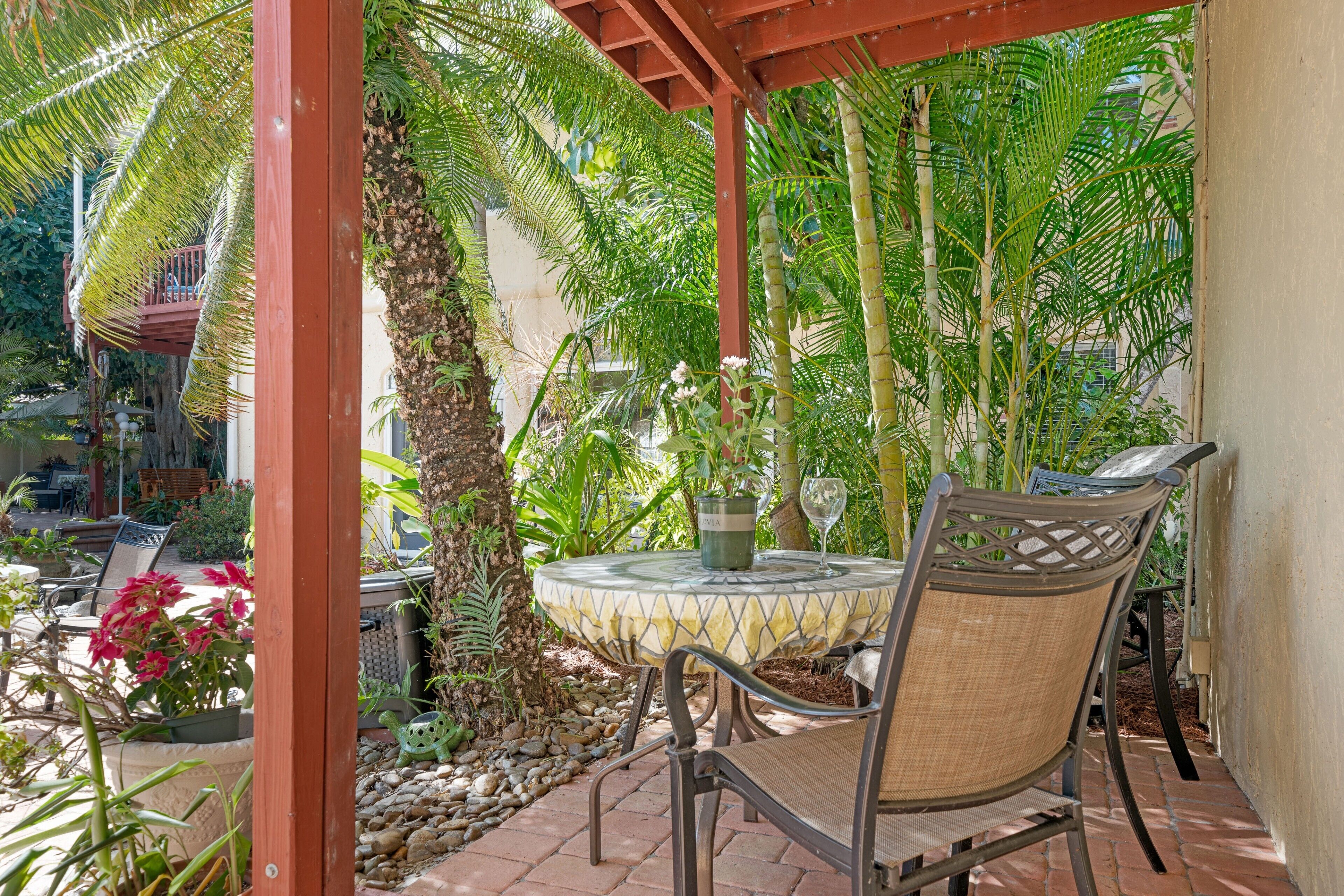A covered outdoor seating area with a table and chairs, surrounded by lush greenery and a variety of plants.