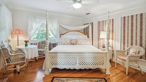 Un dormitorio con cama con dosel, ventilador de techo, ventana con cortinas, una mesita con lámpara y dos sillas de mimbre.