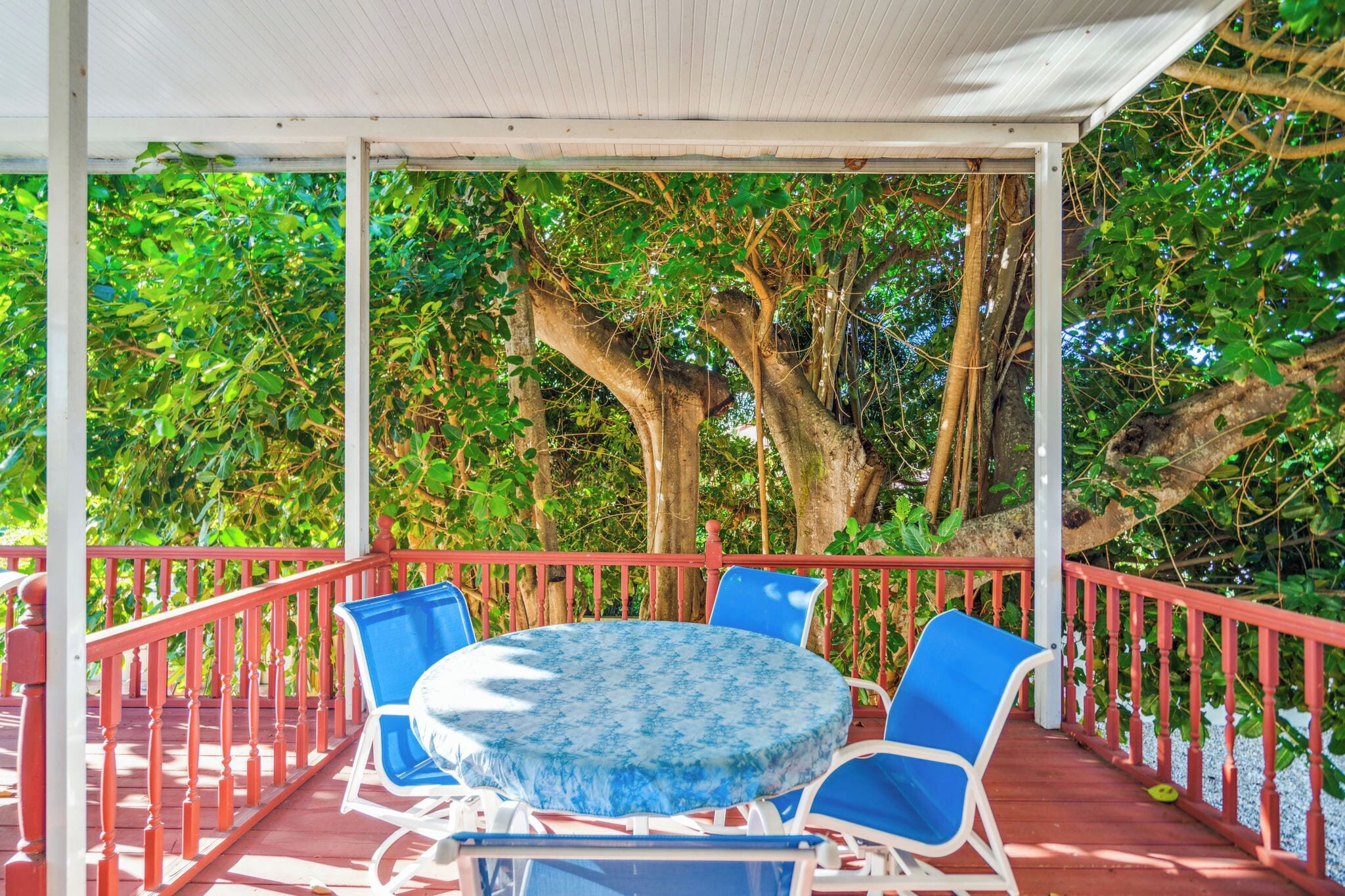 Un patio exterior con una mesa redonda y sillas azules, rodeado por una barandilla roja y árboles.