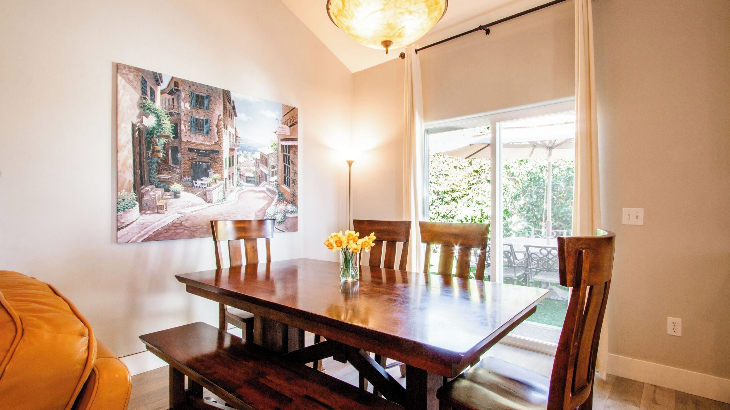 A dining area with a wooden table, chairs, and a vase of flowers. There are two paintings on the wall and a window with a view of greenery.