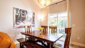 Uma área de refeições com uma mesa de madeira, cadeiras e um vaso de flores. Há duas pinturas na parede e uma janela com vista para o verde.
