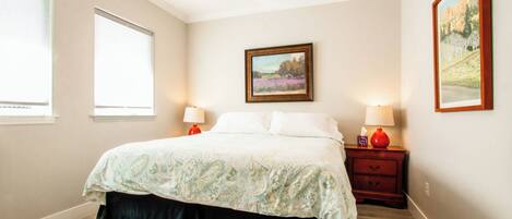 A bedroom with a bed, two bedside tables with lamps, a window, and a painting on the wall.
