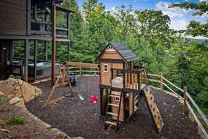 A wooden playhouse with a slide and climbing wall, surrounded by a deck and a hot tub. - Raccoon Ridge - Mountain View | Pet Friendly | Hot Tub | Game Room | Firepit (Blue Ridge)