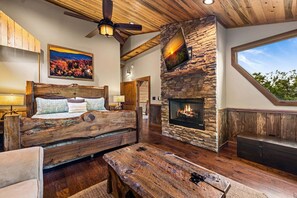 A rustic interior with a stone fireplace, wooden ceiling beams, a bed with a wooden headboard, and a large window. - Raccoon Ridge - Mountain View | Pet Friendly | Hot Tub | Game Room | Firepit (Blue Ridge)