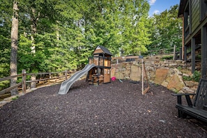 A playground with a wooden slide and a playhouse, surrounded by trees and a stone wall. - Raccoon Ridge - Mountain View | Pet Friendly | Hot Tub | Game Room | Firepit (Blue Ridge)