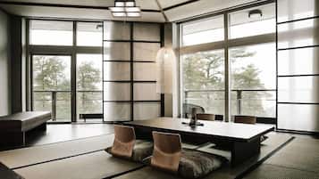 A modern interior with a large dining table, cushioned seating, and large windows offering a view of trees.