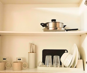 A kitchen with a pot on the upper shelf, a cutting board with plates and glasses on the lower shelf, and a set of mugs on the countertop. - The Executive Suites - Shell Residences Manila 1BR Condo near MOA NAIA Ikea (Pasay City)