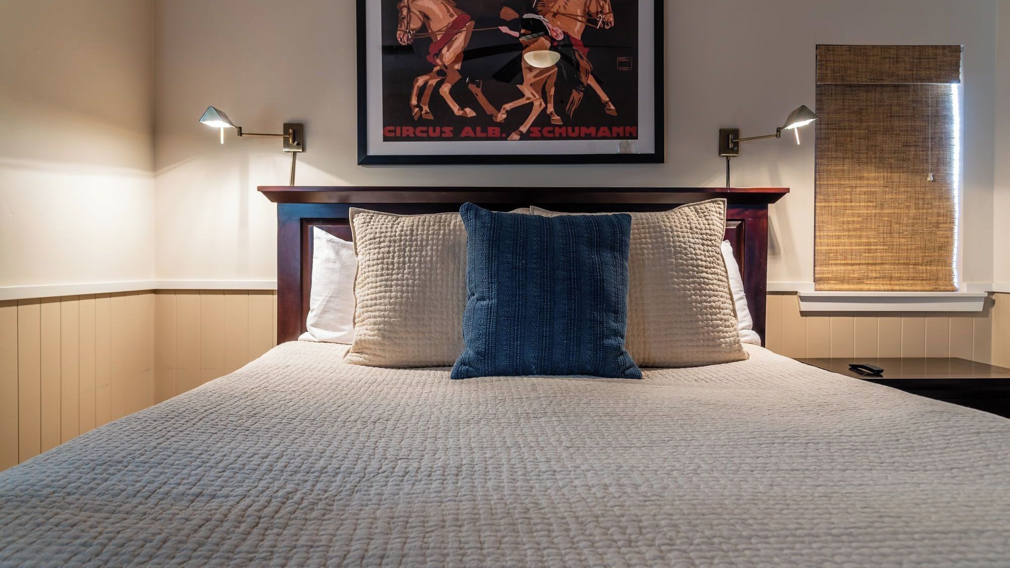 A neatly made bed with a textured white bedspread and two pillows, one blue and one white, in a room with a framed poster on the wall and a window with a blind.