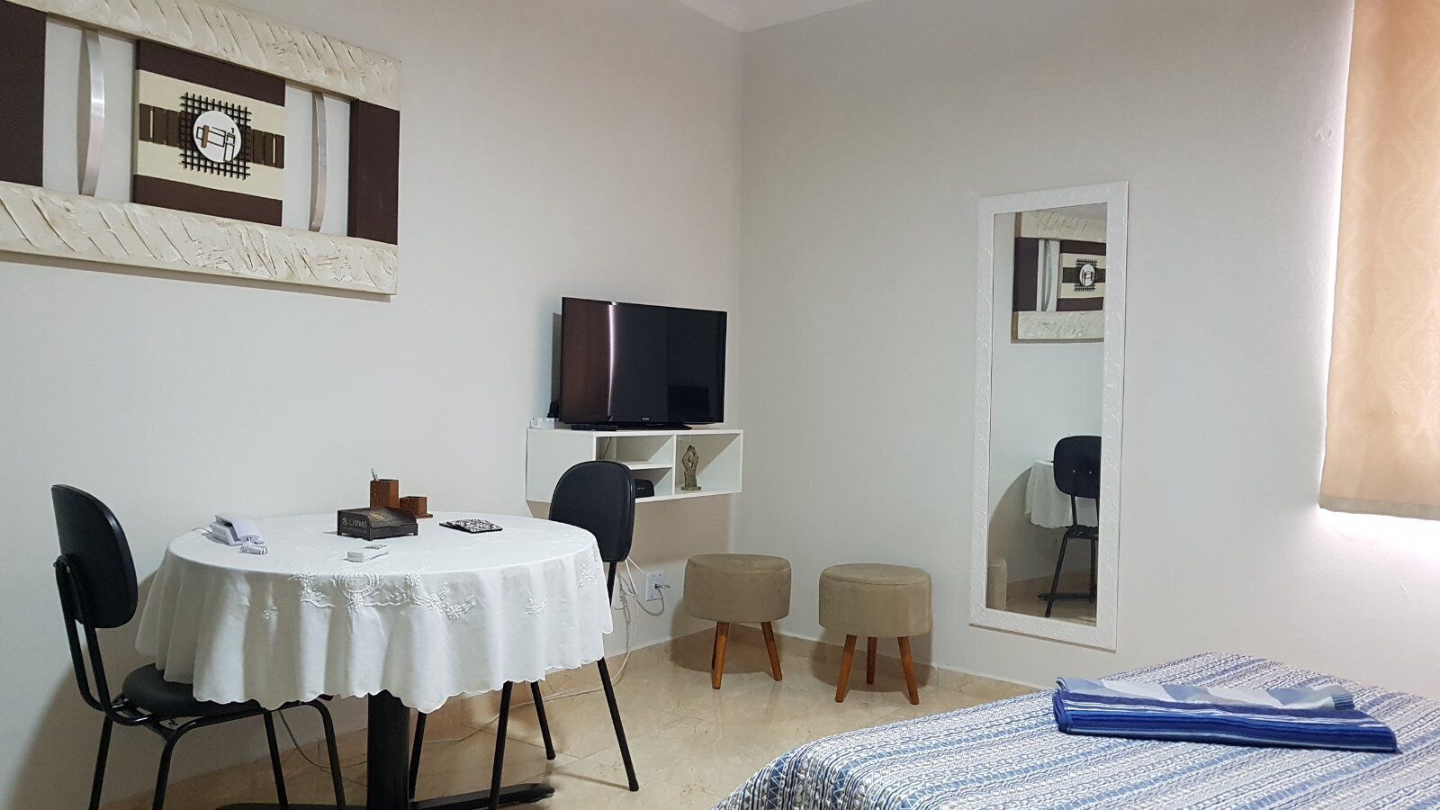 A hotel room with a round dining table, two chairs, a television on a stand, a small stool, a bed with a patterned blanket, and a window with a view.