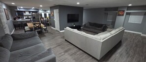 A modern living room with a grey sofa, a dining area with a wooden table and chairs, and a kitchen with a refrigerator and stove.