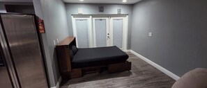 A compact bedroom with a bed, a built-in bench, a refrigerator, and a set of sliding doors.