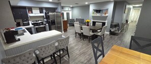 A modern kitchen with stainless steel appliances, a dining area with a wooden table and upholstered chairs, and a grey tiled backsplash.